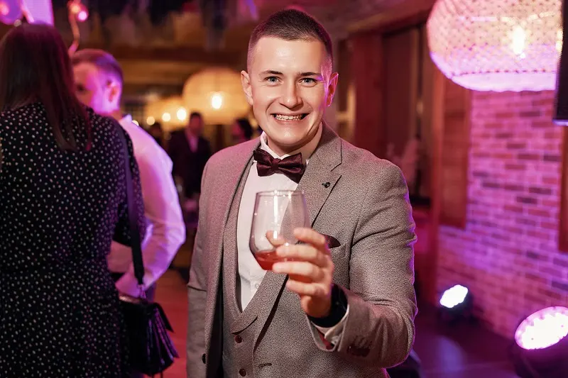 Man in a beige suit and bow tie holding a drink under magenta party lighting