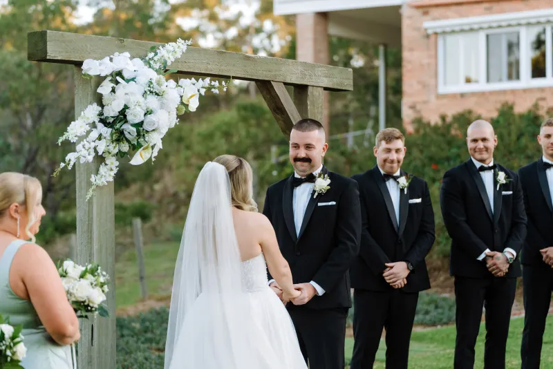 Bride at the altar under a white floral arch, groom and groomsmen turned towards her