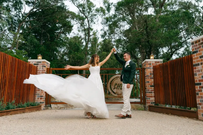 Bride swinging her gown wide at a brick and timber gate while the groom holds her hand