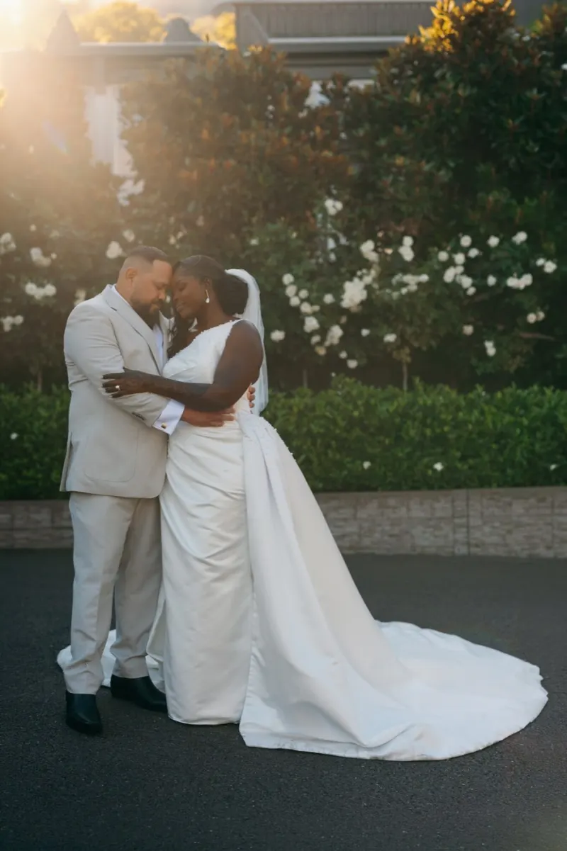 Couple embracing forehead to forehead on a driveway, a backlit garden of white roses behind